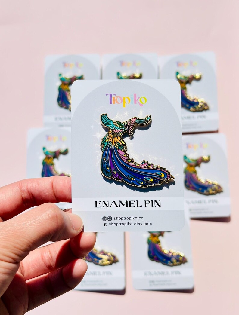 Puede incluir: Un pin de esmalte colorido con un p&aacute;jaro estilizado con una cola larga y fluida. El pin est&aacute; en una tarjeta blanca con el texto "Tropiko Enamel Pin" y la direcci&oacute;n del sitio web "shoptropiko.co".