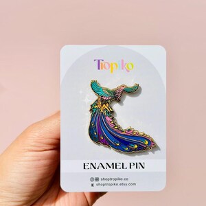 Puede incluir: Un pin de esmalte colorido que presenta un p&aacute;jaro estilizado con una cola larga y fluida. El p&aacute;jaro est&aacute; representado en tonos de rosa, azul, verde y amarillo. El pin est&aacute; en una tarjeta blanca con el texto "Tropiko" y "Enamel Pin" impreso en ella.