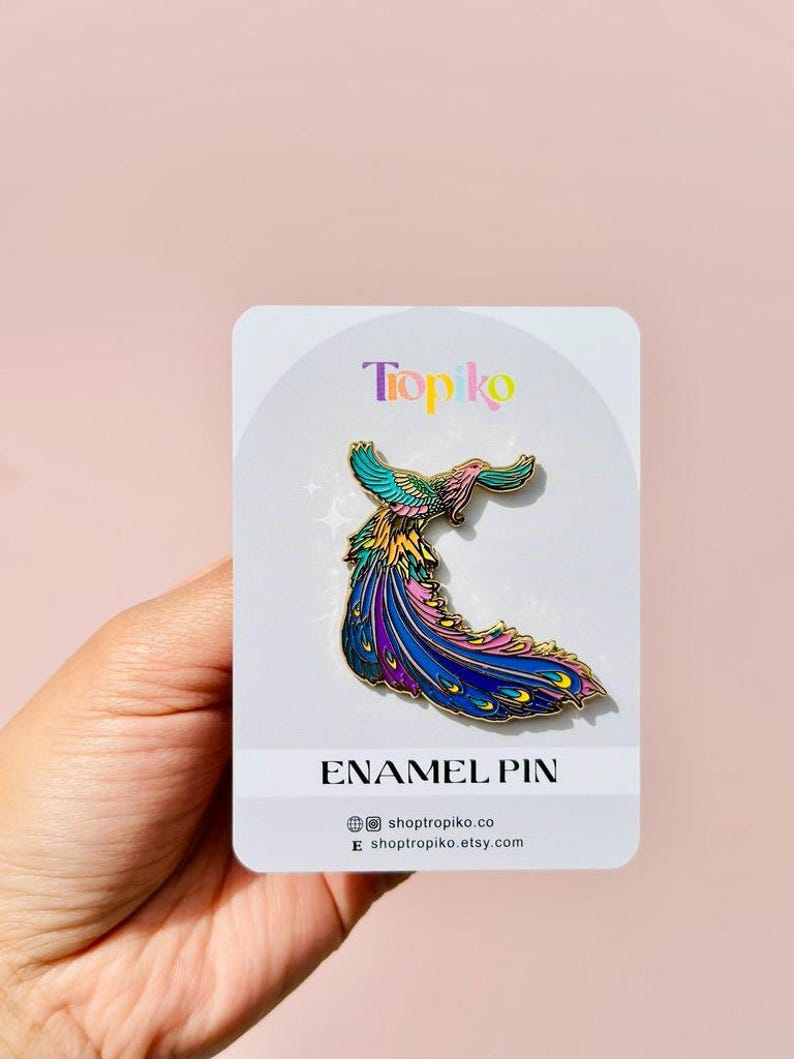 Puede incluir: Un pin de esmalte colorido que presenta un p&aacute;jaro estilizado con una cola larga y fluida. El p&aacute;jaro est&aacute; representado en tonos de rosa, azul, verde y amarillo. El pin est&aacute; en una tarjeta blanca con el texto "Tropiko" y "Enamel Pin" impreso en ella.