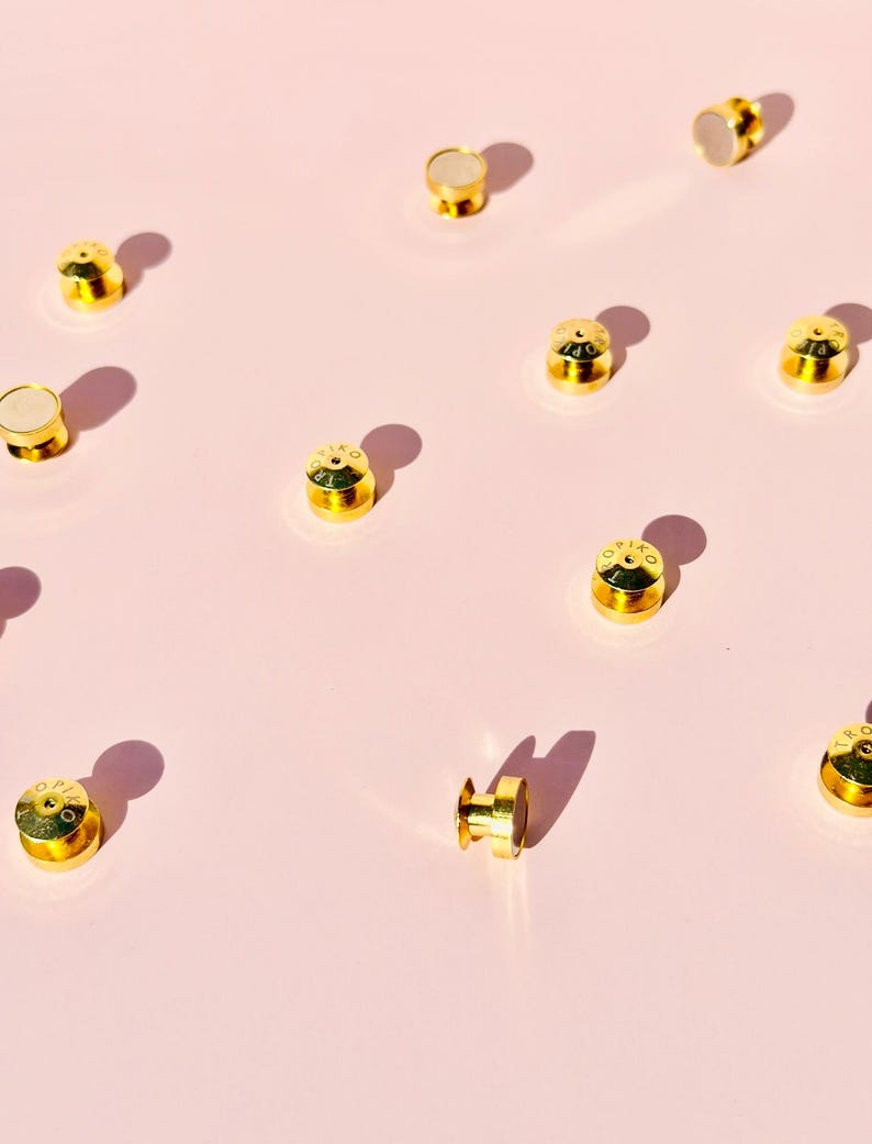 Puede incluir: Una colecci&oacute;n de broches met&aacute;licos de color dorado con un centro blanco. Los broches est&aacute;n dispersos sobre una superficie rosa.