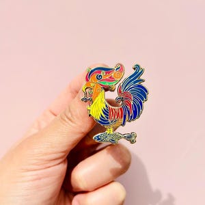 May include: A colorful enamel pin featuring a rooster with a fish in its beak. The rooster is standing on a fish with its tail feathers spread out. The pin is gold-toned and has a detailed design.