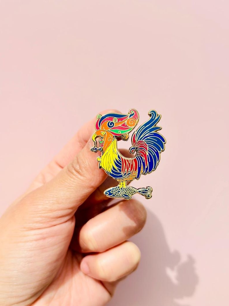 May include: A colorful enamel pin featuring a rooster with a fish in its beak. The rooster is standing on a fish with its tail feathers spread out. The pin is gold-toned and has a detailed design.