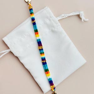 May include: A colorful beaded bracelet with a rainbow pattern and gold accents. The bracelet is on a white drawstring bag with the word "Audrey" on the tag.