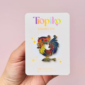 May include: A colorful enamel pin featuring a rooster with a fish in its beak. The rooster is depicted in a vibrant red, yellow, blue, and green color scheme. The pin is on a white card with the text "Tropiko Enamel Pin" and social media handles.