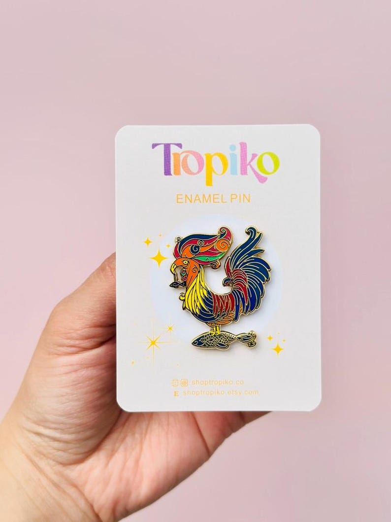 May include: A colorful enamel pin featuring a rooster with a fish in its beak. The rooster is depicted in a vibrant red, yellow, blue, and green color scheme. The pin is on a white card with the text "Tropiko Enamel Pin" and social media handles.