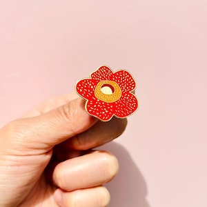 Op de afbeelding: Een rode en gele emaille pin in de vorm van een bloem. De pin wordt in een hand gehouden.