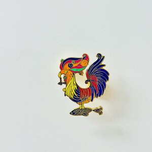 May include: A gold-colored enamel pin featuring a colorful rooster standing on a fish. The rooster has a red, yellow, and blue body with intricate details. The fish is blue and black.