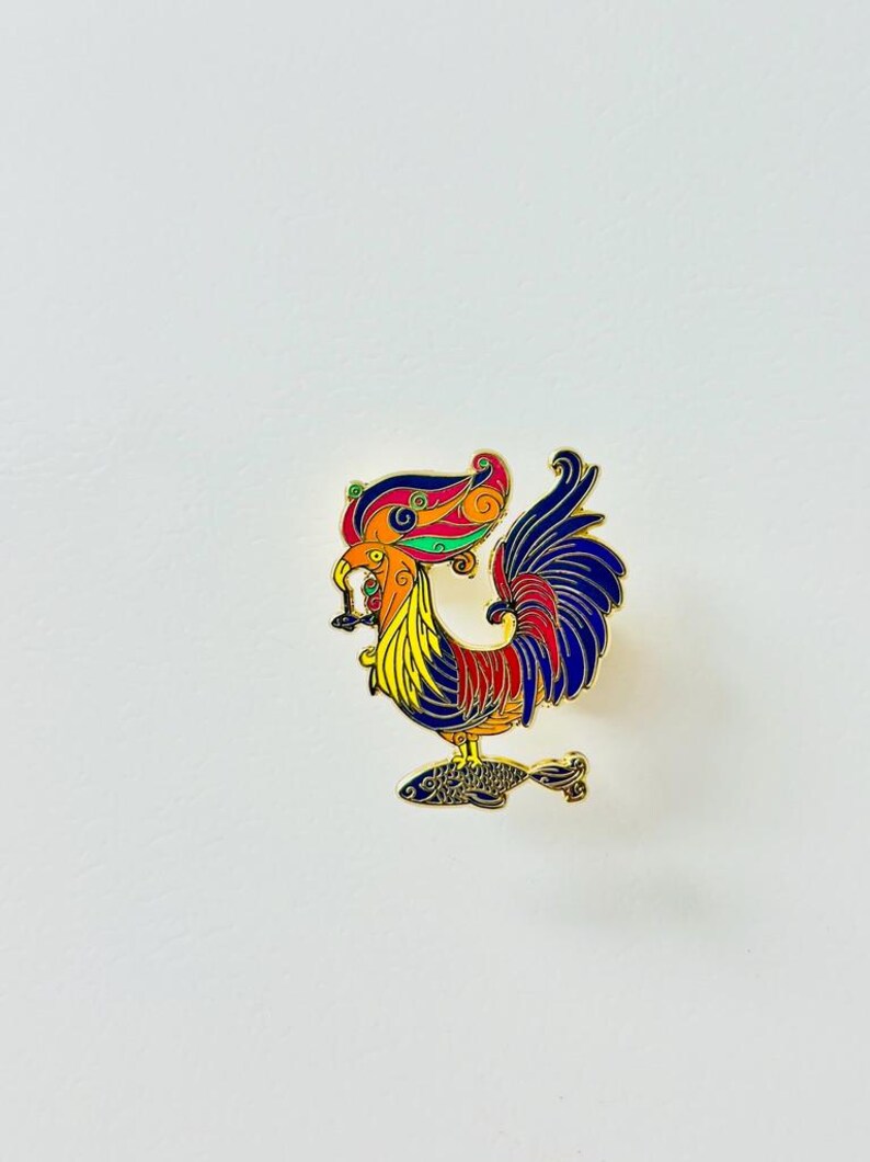 May include: A gold-colored enamel pin featuring a colorful rooster standing on a fish. The rooster has a red, yellow, and blue body with intricate details. The fish is blue and black.