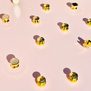 May include: A collection of 12 gold-colored metal bottle stoppers with various designs and lettering. The stoppers are arranged on a pink surface.