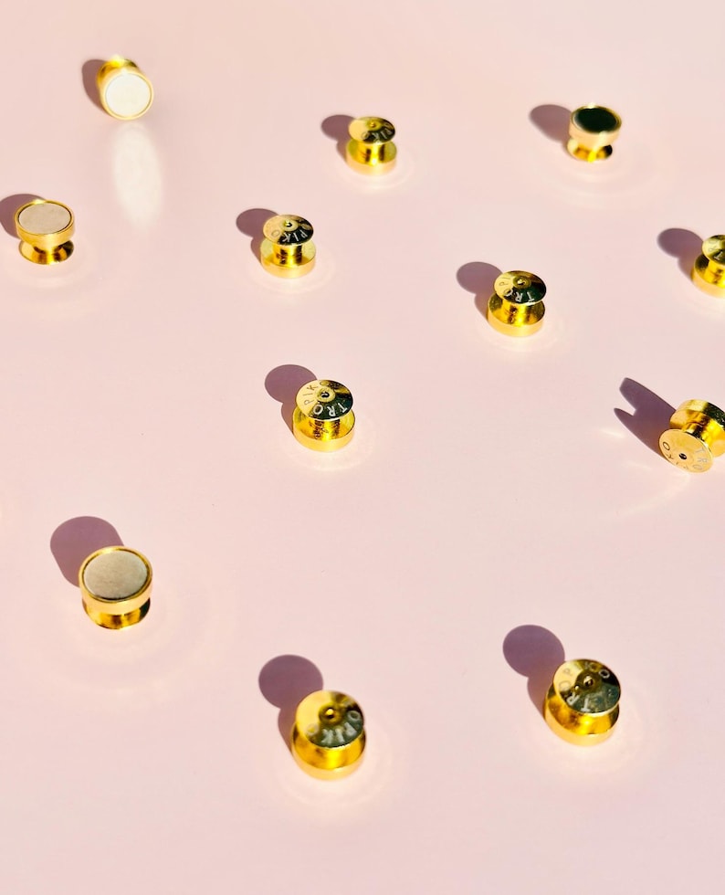 May include: A collection of 12 gold-colored metal bottle stoppers with various designs and lettering. The stoppers are arranged on a pink surface.