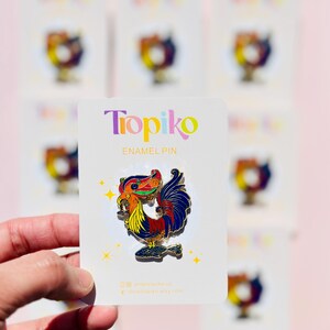 May include: A colorful enamel pin featuring a rooster with a detailed design. The rooster is holding a fish in its beak and has a bright red, yellow, blue, and green color scheme. The pin is on a white card with the text "Topiko Enamel Pin" and the shop's website address.