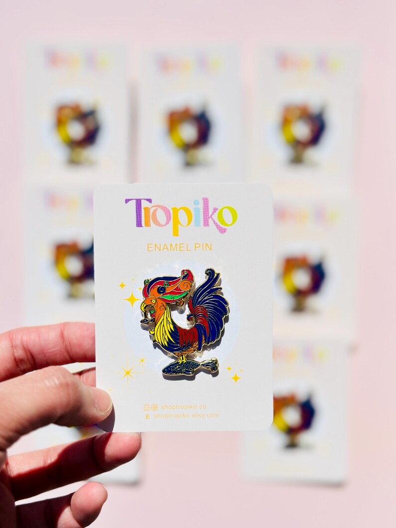 May include: A colorful enamel pin featuring a rooster with a detailed design. The rooster is holding a fish in its beak and has a bright red, yellow, blue, and green color scheme. The pin is on a white card with the text "Topiko Enamel Pin" and the shop's website address.