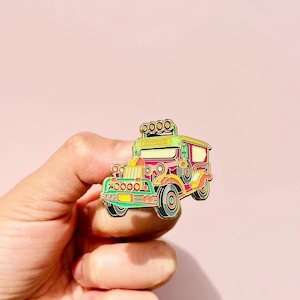 Op de afbeelding: Een groene en rode jeepney-emaille pin met gouden accenten. De jeepney heeft een geel dak en een wit kenteken met zwarte tekst. De pin is een populair souvenir uit de Filippijnen.