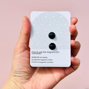 Puede incluir: Dos convertidores de pines magn&eacute;ticos negros en una tarjeta blanca con instrucciones sobre c&oacute;mo usarlos. Las instrucciones dicen: "C&oacute;mo usar el convertidor de pines magn&eacute;tico: RETIRAR las espaldas de los pines DESLIZAR los cierres magn&eacute;ticos PEGAR en una superficie magn&eacute;tica"