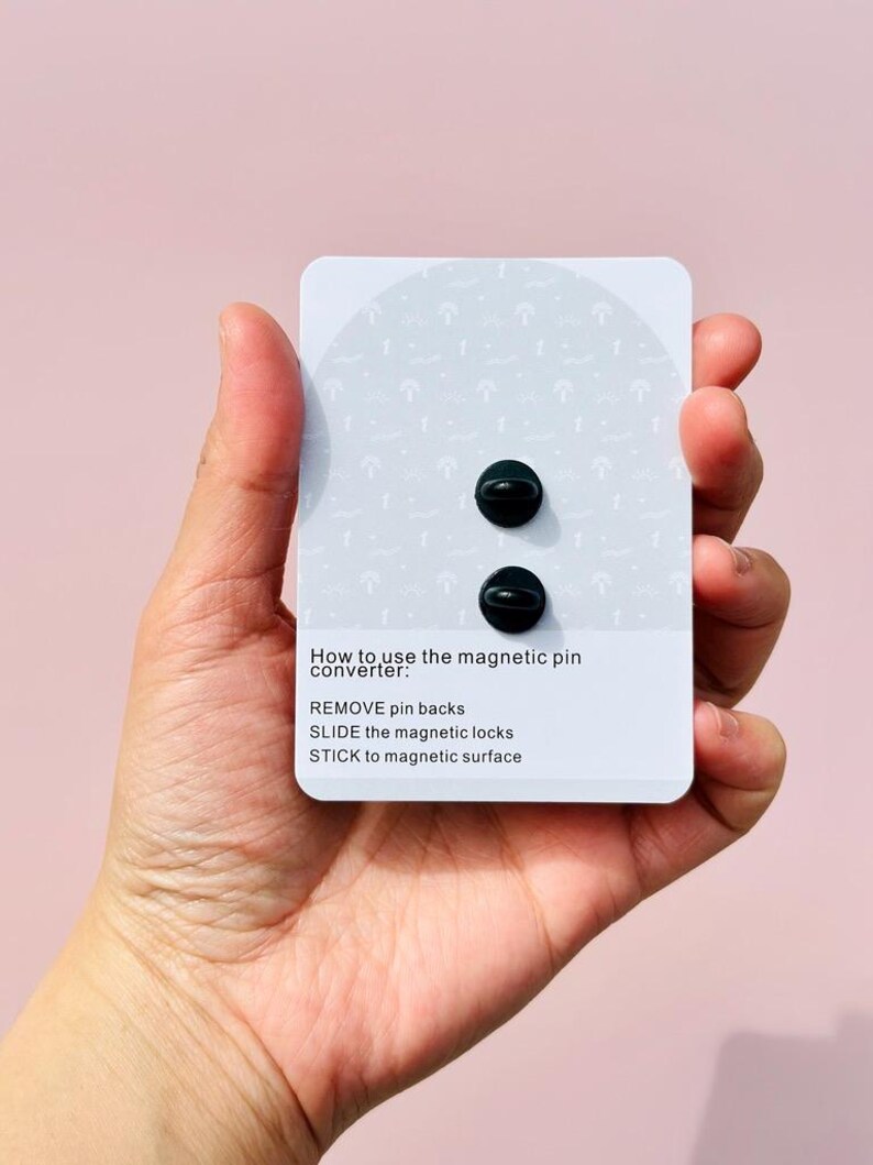 Puede incluir: Dos convertidores de pines magn&eacute;ticos negros en una tarjeta blanca con instrucciones sobre c&oacute;mo usarlos. Las instrucciones dicen: "C&oacute;mo usar el convertidor de pines magn&eacute;tico: RETIRAR las espaldas de los pines DESLIZAR los cierres magn&eacute;ticos PEGAR en una superficie magn&eacute;tica"