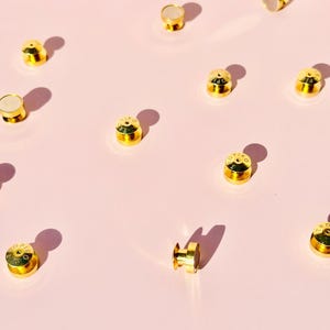 May include: A collection of gold-colored metal snaps with a white center. The snaps are scattered on a pink surface.