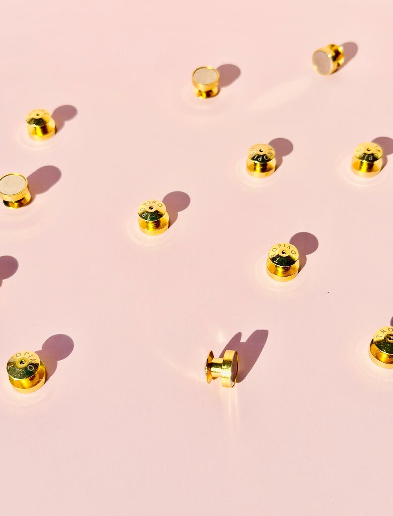 May include: A collection of gold-colored metal snaps with a white center. The snaps are scattered on a pink surface.