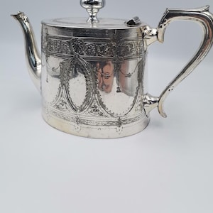 May include: A silver teapot with an ornate design. The teapot has a lid with a knob and a spout. The handle is curved and has a decorative design.