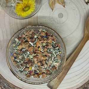 Op de afbeelding: Een glazen kom gevuld met een mengsel van gedroogde kruiden, vruchten en bloemen. Het mengsel bevat stukjes gedroogde appel, citroen en kamillebloemen.