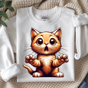 May include: White crewneck sweatshirt featuring a cartoon orange cat with large eyes and an open mouth. The cat has its paws outstretched. The sweatshirt is made of a soft material.