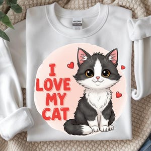 Peut inclure: Sweat-shirt blanc avec un chat de dessin animé noir et blanc, de grands yeux et un nez rose. Le texte "I LOVE MY CAT" est en rouge. De petits coeurs rouges sont dispersés autour du chat. Le sweat-shirt est en matière douce.