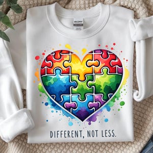 Könnte beinhalten: Weißes Sweatshirt mit einem herzförmigen Puzzlemotiv in bunten Farben. Die Puzzleteile sind rot, gelb, blau und grün. Der Text "DIFFERENT, NOT LESS" ist darunter aufgedruckt. Aquarellspritzer umgeben das Herz.