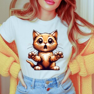 May include: White t-shirt featuring a cartoon orange cat with large eyes and an open mouth, as if surprised. The cat's paws are outstretched. The shirt is paired with blue jeans and a yellow sweater.