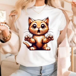 May include: White t-shirt featuring a cartoon orange kitten with large eyes and an open mouth, as if surprised. The kitten's paws are outstretched. The design is centered on the shirt.