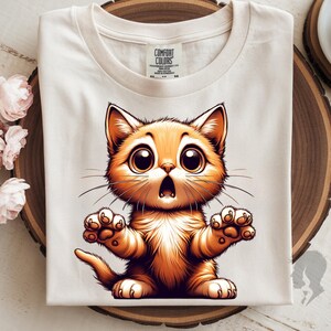 May include: Cream-colored t-shirt featuring a cartoon orange cat with large, expressive eyes and an open mouth. The cat's paws are outstretched, and the shirt has a Comfort Colors tag.