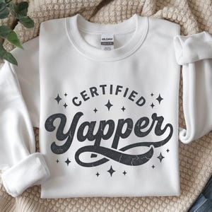May include: White crewneck sweatshirt with the text "CERTIFIED YAPPER" in a distressed black font, surrounded by star graphics. The sweatshirt is made of a soft material and is suitable for casual wear.