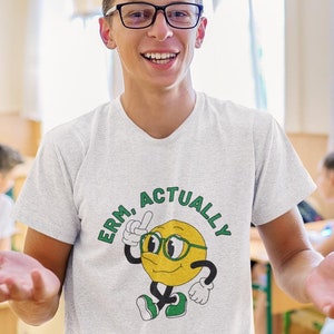 Puede incluir: Camiseta gris claro con el texto "ERM, ACTUALLY" y un emoji de dibujos animados con gafas. El emoji es amarillo con contornos negros y zapatos verdes. La persona lleva gafas y sonríe.