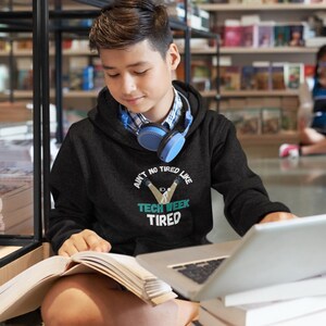 Op de afbeelding: Een zwarte hoodie met de tekst "Ain't no tired like Tech Week Tired" in wit en teal. De hoodie wordt gedragen met een blauwe koptelefoon en een geruit overhemd. Een laptop en een boek staan op de voorgrond.