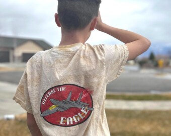 Puede incluir: Camiseta beige de manga corta con un gráfico de un avión de combate dentro de un óvalo rojo, con el texto "RELEASE THE EAGLE". La camiseta la lleva una persona con el pelo corto castaño.