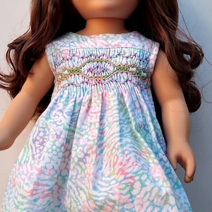 May include: A doll wearing a white dress with a colorful floral pattern and green and purple embroidery. The doll has long brown hair and green eyes. She is wearing brown boots.
