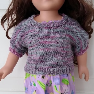 Könnte beinhalten: Eine Puppe mit braunen Haaren und grünen Augen, die einen handgestrickten grau-violett-rosa Pullover und einen lavendelfarbenen Rock mit Blumen- und Vogelmuster trägt. Die Puppe steht vor einem weißen Hintergrund.