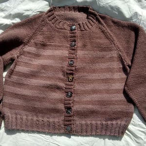 以下が含まれることがあります： 茶色のボタン付きカーディガン。リブ編みの襟と袖口があり、茶色の濃淡の水平ストライプ模様です。濃い色のボタンで留められています。手編みの、居心地の良い衣服です。