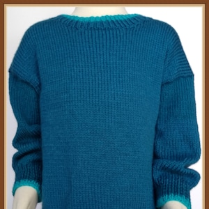 Op de afbeelding: Een tealblauwe gebreide trui met een lichtblauwe rand rond de hals en zoom. De trui is op een mannequin die een paars-witte geruite rok draagt.