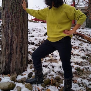 Könnte beinhalten: Eine Person in einem leuchtend gelben Pullover und einer schwarzen Hose steht in einer schneebedeckten Landschaft. Sie trägt schwarze Stiefel und hat ihre Hand an einem Baumstamm.