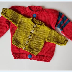 Puede incluir: Dos cárdigans de punto, uno rojo con rayas azules en las mangas y otro verde oliva. Ambos cárdigans tienen botones de madera.