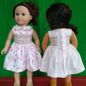 May include: Two dolls wearing white dresses with pink leaf patterns and green and pink embroidered bodices. The dresses have a Peter Pan collar and button closures in the back. One doll has brown hair and gold glitter shoes, the other has black hair and yellow shoes.