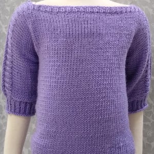 Puede incluir: Un suéter de punto lavanda con cuello barco y mangas cortas. Las mangas tienen un diseño texturizado. El suéter se muestra en un maniquí. El dobladillo inferior y el escote tienen un diseño acanalado.