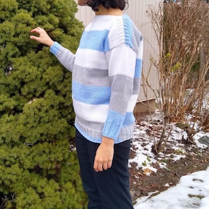 May include: A knitted sweater with horizontal stripes in light blue, white, and grey. The sweater has long sleeves and a crew neck. The person is wearing black trousers and black boots. The background includes a green bush and snow.