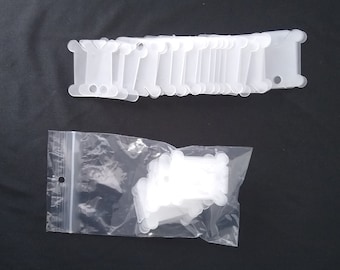 May include: A collection of white plastic bobbins, designed for thread storage, are displayed against a dark background. Some bobbins are neatly arranged, while others are contained within a clear plastic bag. The bobbins have a distinctive shape with rounded edges and holes.