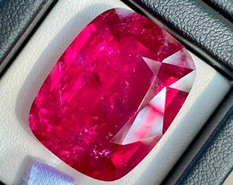 Op de afbeelding: Een grote, kussengeslepen, levendige roze-rode edelsteen met meerdere facetten, gepresenteerd in een zwarte presentatiedoos met een witte binnenvoering en een klein wit stoffen lipje aan de onderkant van de zetting van de steen.