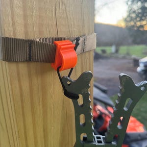 May include: A close-up of a black and gray tree stand with a strap and orange buckle attached to a wooden post. The stand has a serrated metal design and is used for hunting.