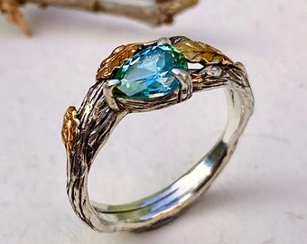 May include: A ring featuring a pear-shaped blue-green gemstone set in a band with a textured, bark-like finish. The band is primarily silver-toned with gold-toned leaf accents flanking the central stone, creating a detailed, organic appearance.