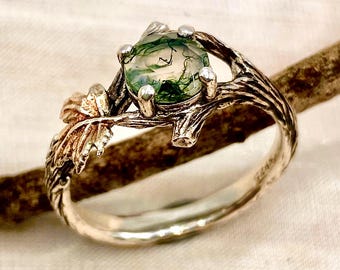 Op de afbeelding: Een ring met een ronde, doorschijnende groene mosagaatsteen in een zilveren band die is ontworpen als een getextureerde boomtak met kleine blaadjes. Het metaal heeft een gepolijste afwerking en de steen vertoont natuurlijke donkergroene insluitingen.