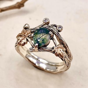 May include: A silver ring with a nature-inspired design. The ring features a pear-shaped, green gemstone at the center, surrounded by silver branches and gold-toned leaves. Two small, clear gemstones are set on the ring's band.