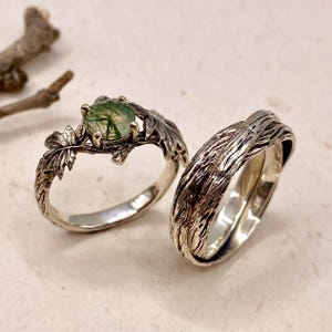 May include: A silver ring featuring a round moss agate stone set within a band designed to resemble textured tree branches and leaves, paired with a second silver band that has a deeply grooved, bark-like texture across its entire surface.