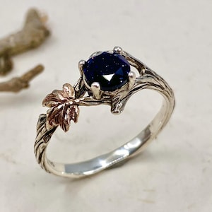 May include: A ring featuring a round, dark blue, glittery gemstone set in a four-prong setting. The band is designed to resemble a textured tree branch with a small, metallic, rose-colored leaf detail extending from the side of the setting.